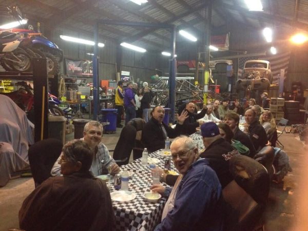 A group of people eating dinner at a long table covered with a checkered tablecloth inside an auto repair shop.