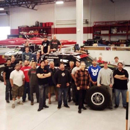 A large group of people posing for a photo in front of a race truck inside a garage.