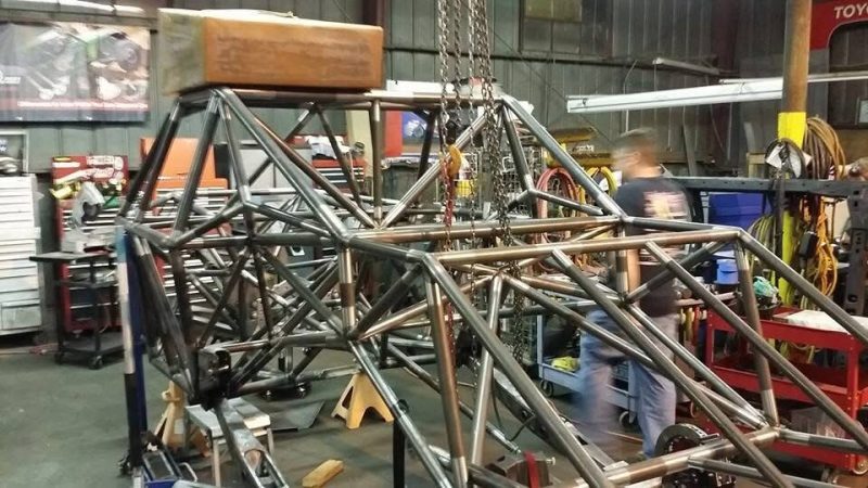 A mechanic working on a custom tubular steel vehicle frame suspended by a chain hoist in a workshop.