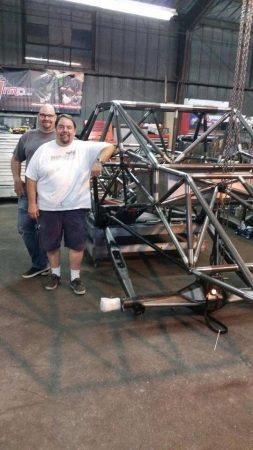 Two men stand next to a large, unfinished tubular metal vehicle frame inside a workshop.