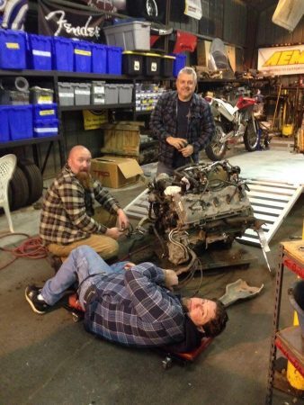 Three men work on an engine sitting on a stand in a garage.