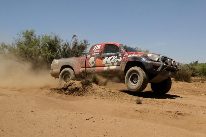 A red and black off-road racing truck numbered 779 jumps over a dirt mound, kicking up a cloud of dust.