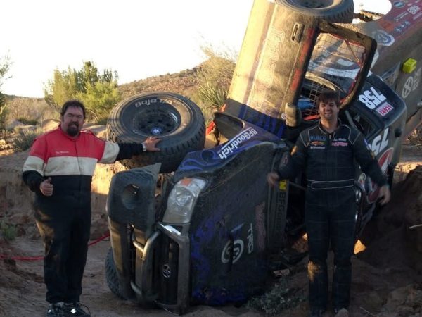 Two men in racing suits stand next to an off-road race truck that has rolled onto its side.