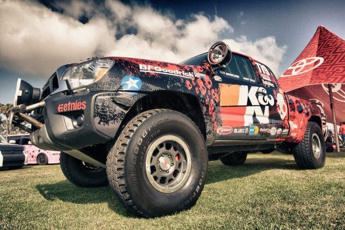A modified Toyota Tacoma off-road race truck with K&N, BFGoodrich, and Etnies branding parked on grass under a cloudy sky.