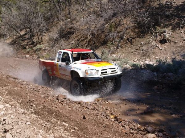 A white and red off-road racing truck splashes through a shallow creek on a dirt trail.