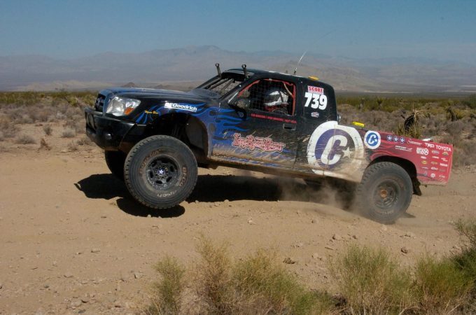 A black off-road racing truck, numbered 739, jumps over a dirt trail in the desert.