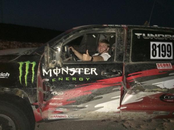 A smiling man gives a thumbs-up from the drivers seat of a muddy, sponsored Toyota Tundra off-road racing truck.