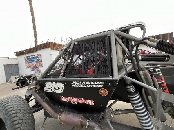 Side view of a black off-road racing vehicle with a roll cage, marked with the number 210 and the names Jach Moncure and Jorge Lameda.