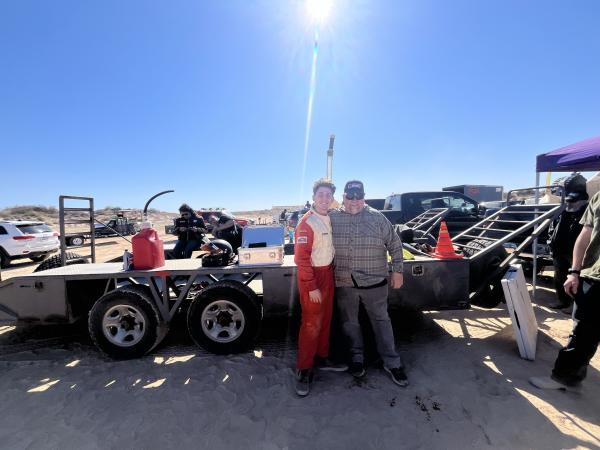 A young man in a red racing suit and an older man in a plaid shirt stand in front of a flatbed trailer on a sandy lot.