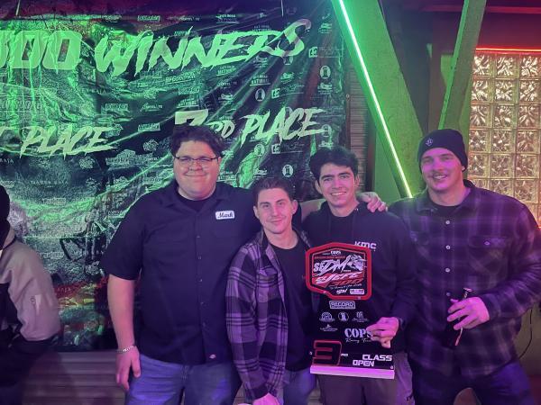 Four men standing in front of a green backdrop for the San Felipe 250 race, with one man holding a third-place trophy.
