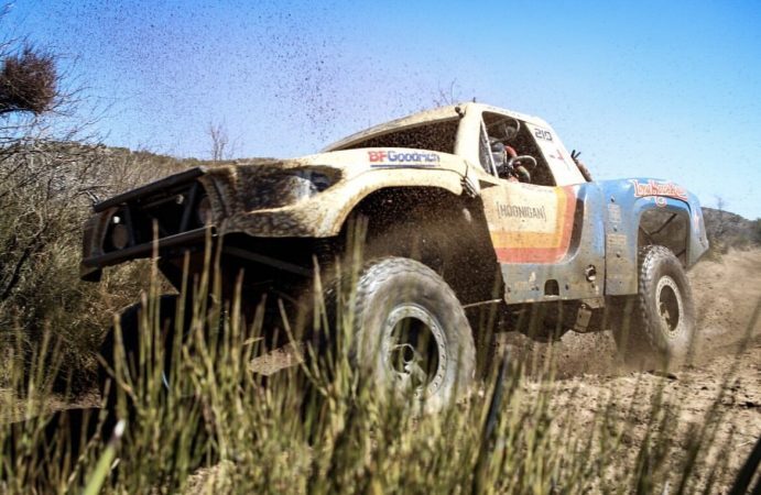 A trophy truck with a blue, yellow, and red paint scheme speeding through a dirt trail, kicking up a cloud of dust.
