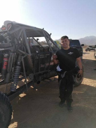 A smiling man standing next to an off-road race car in a desert environment.