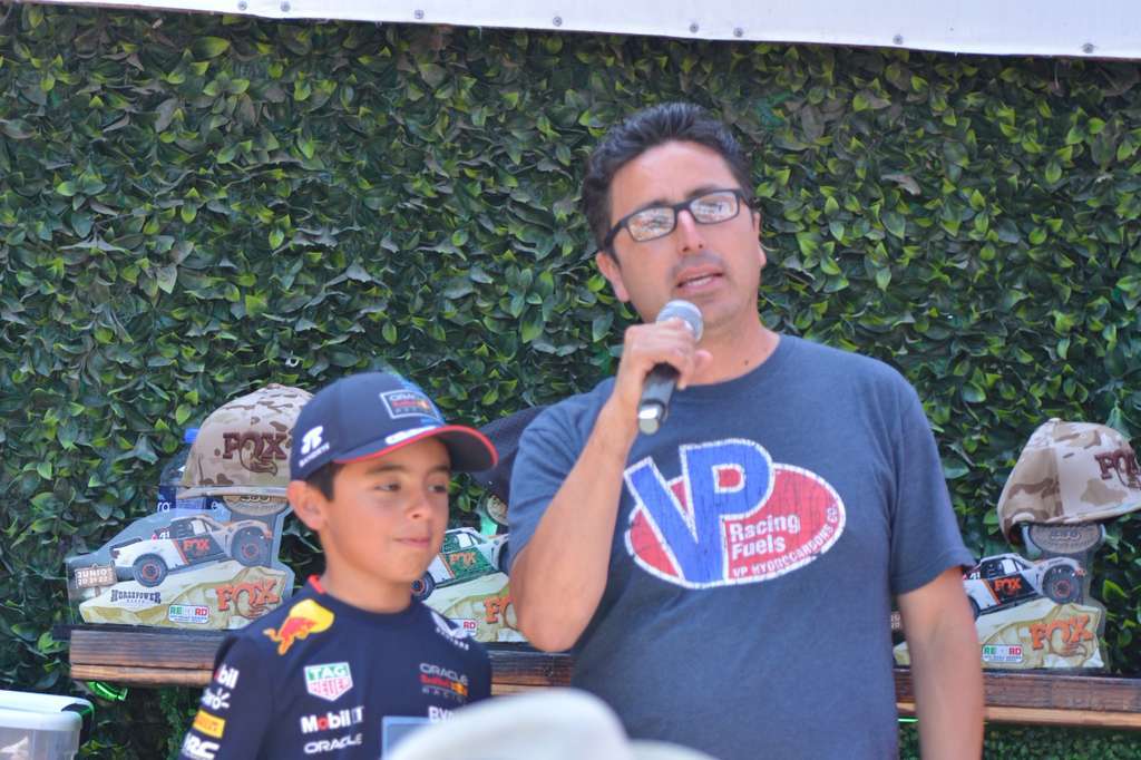 A man speaking into a microphone stands next to a young boy wearing a racing team shirt and cap, in front of a shelf displaying racing-themed toy cars.
