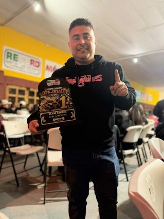 A man in a black hooded sweatshirt holds a first-place off-road racing trophy while pointing upward with his index finger.