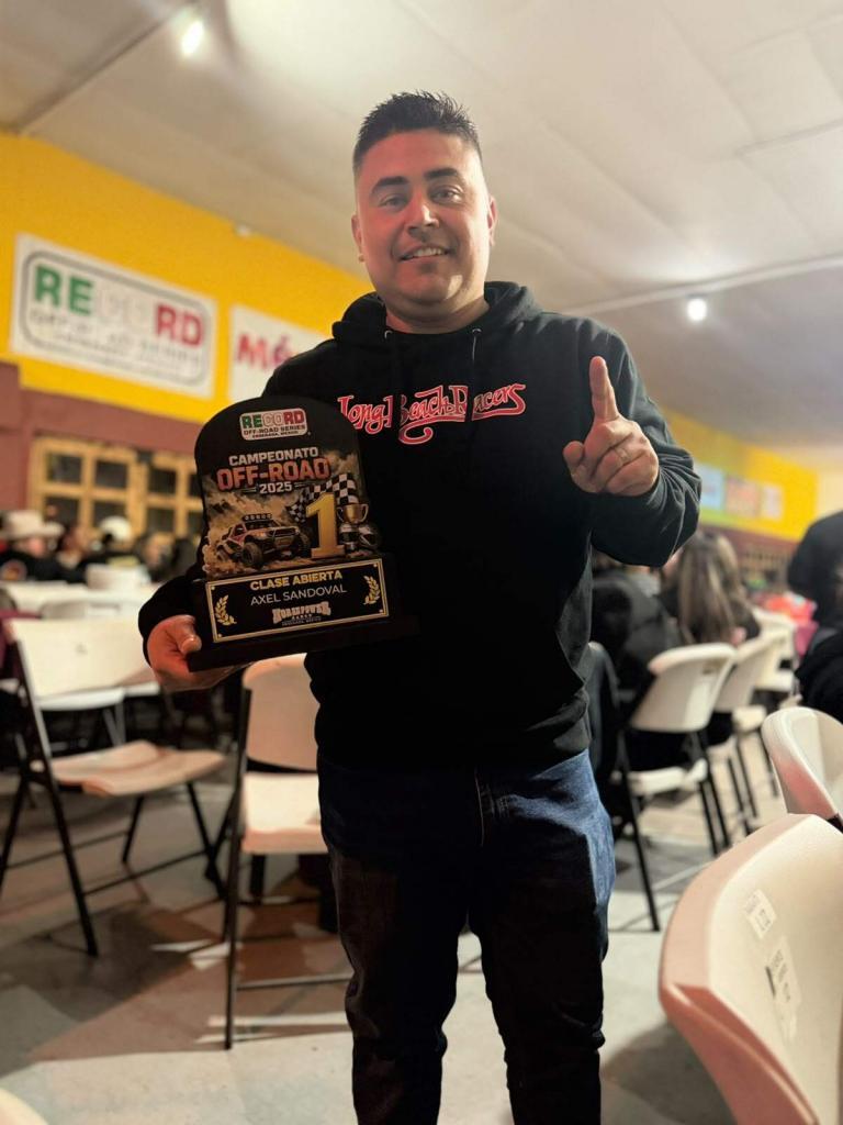 A man in a black hooded sweatshirt holds a first-place off-road racing trophy while pointing upward with his index finger.