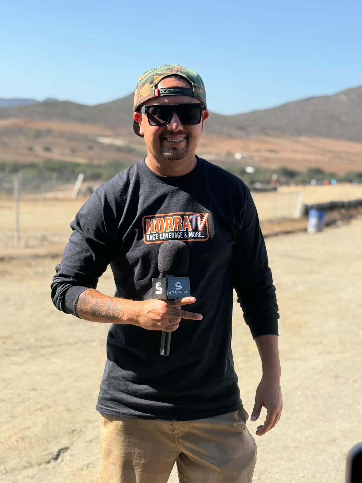 A reporter wearing a black NORRA TV long-sleeve shirt, sunglasses, and a camouflage cap holds a microphone in a desert setting.