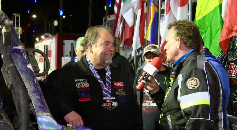 A reporter holding a microphone interviews a man wearing a Baja 500 finisher medal in front of race flags.